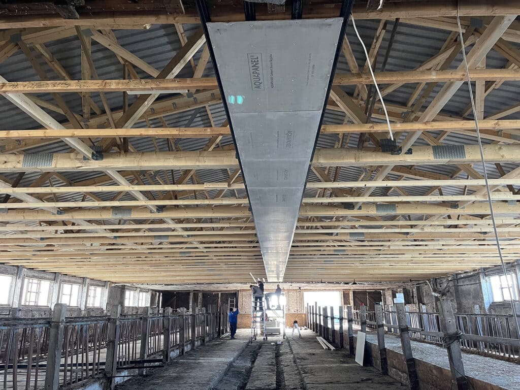 Dachsanierung in einer Gewerbebaustelle: Blick von unten auf eine große grau-schwarze Trapezblech-Dachbahn, gespannt unter Holzdeckenkonstruktion mit Stahlbalken. Arbeiter auf Gerüst bei der Dachneueindeckung.